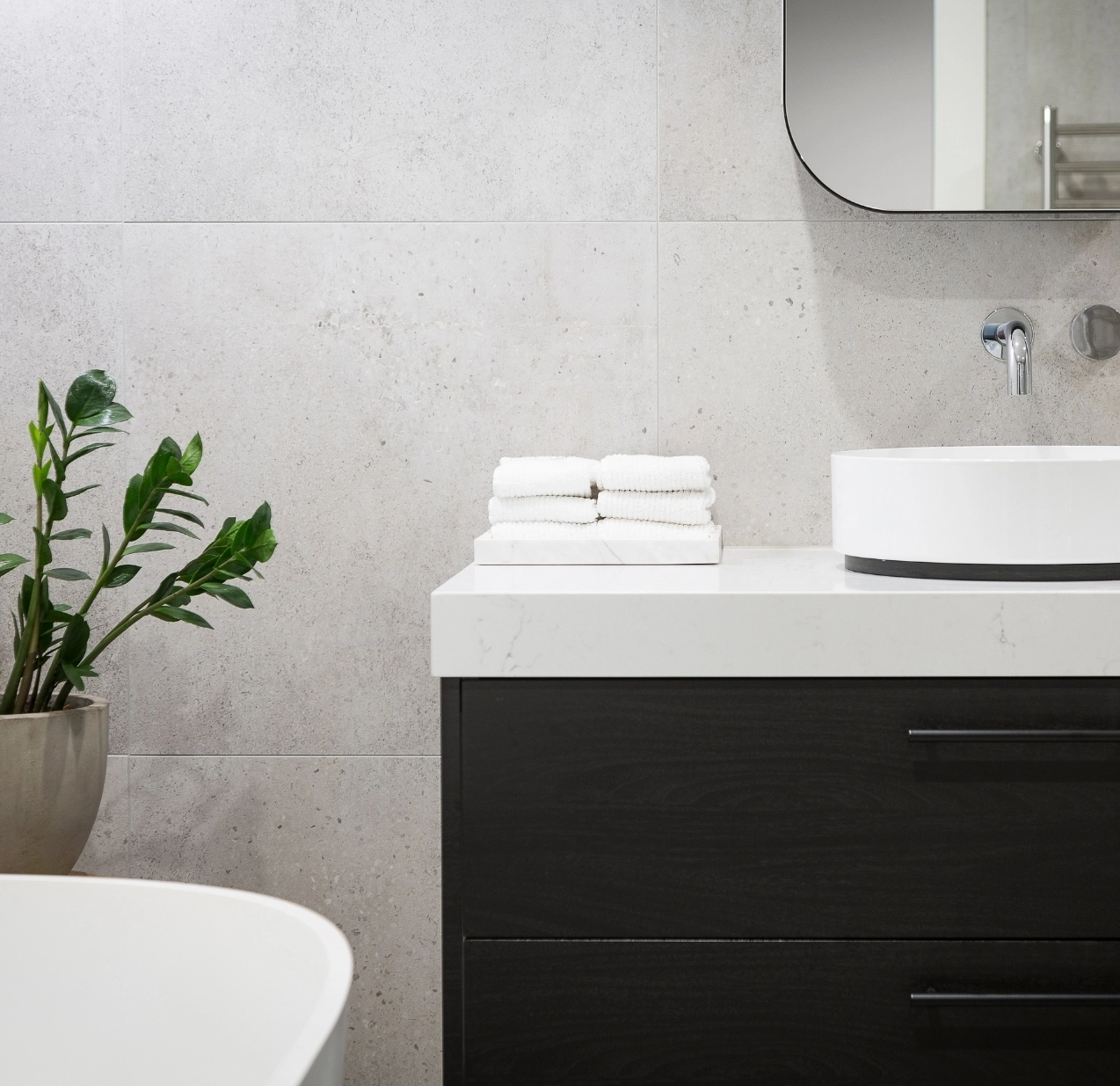Modernes Badezimmer mit dunklem Waschtisch, weißem Aufsatzwaschbecken, grauen Fliesen und Zimmerpflanze.