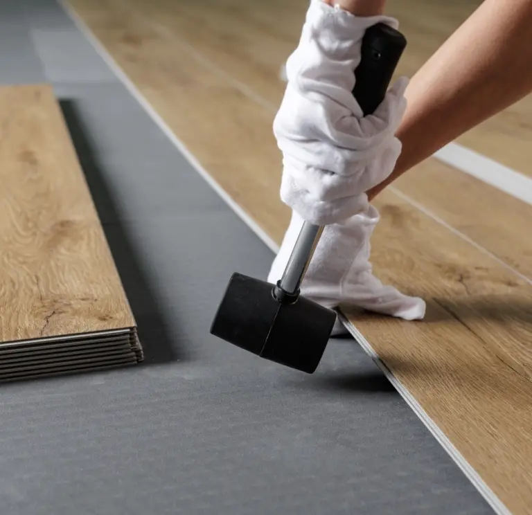 Person verlegt Holzoptik-Laminat- oder Vinylboden mit Gummihammer auf grauem Untergrund.