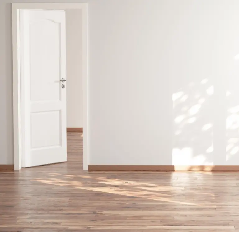 Helles, leeres Zimmer mit Holzboden, weißer Wand, offener Tür und Sonnenlicht, das Blattschatten wirft.