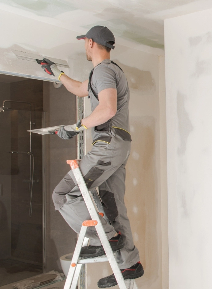 Handwerker in grauer Arbeitskleidung spachtelt Wand und Decke auf einer Trittleiter während einer Renovierung.